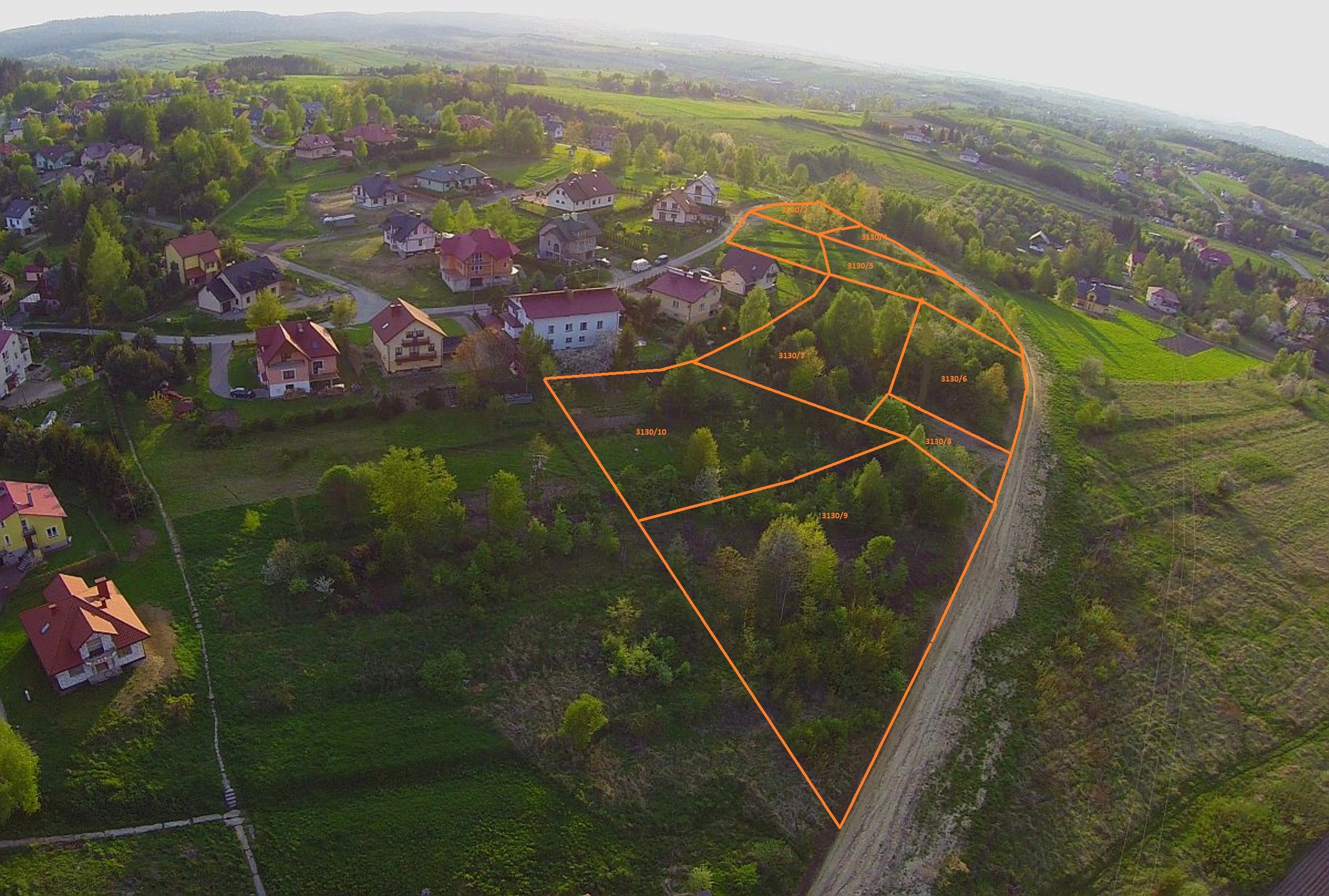 Zdjęcie lotnicze wiejskiego krajobrazu z zaznaczonymi pomarańczowymi liniami granic działek gruntu. Widoczne domy i zieleń.